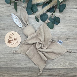 May include: A beige muslin baby bunny lovey with knotted corners and a floral print on the ears. The lovey is on a wooden surface with a wooden sign that says "hello little one" with a star and shooting stars.