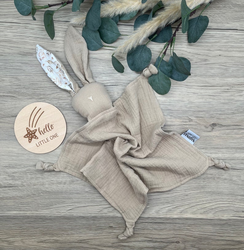 May include: A beige muslin baby bunny lovey with knotted corners and a floral print on the ears. The lovey is on a wooden surface with a wooden sign that says "hello little one" with a star and shooting stars.