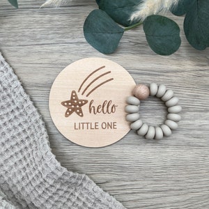 May include: A wooden round sign with a shooting star and the text "hello little one". A gray silicone teething ring is next to the sign.