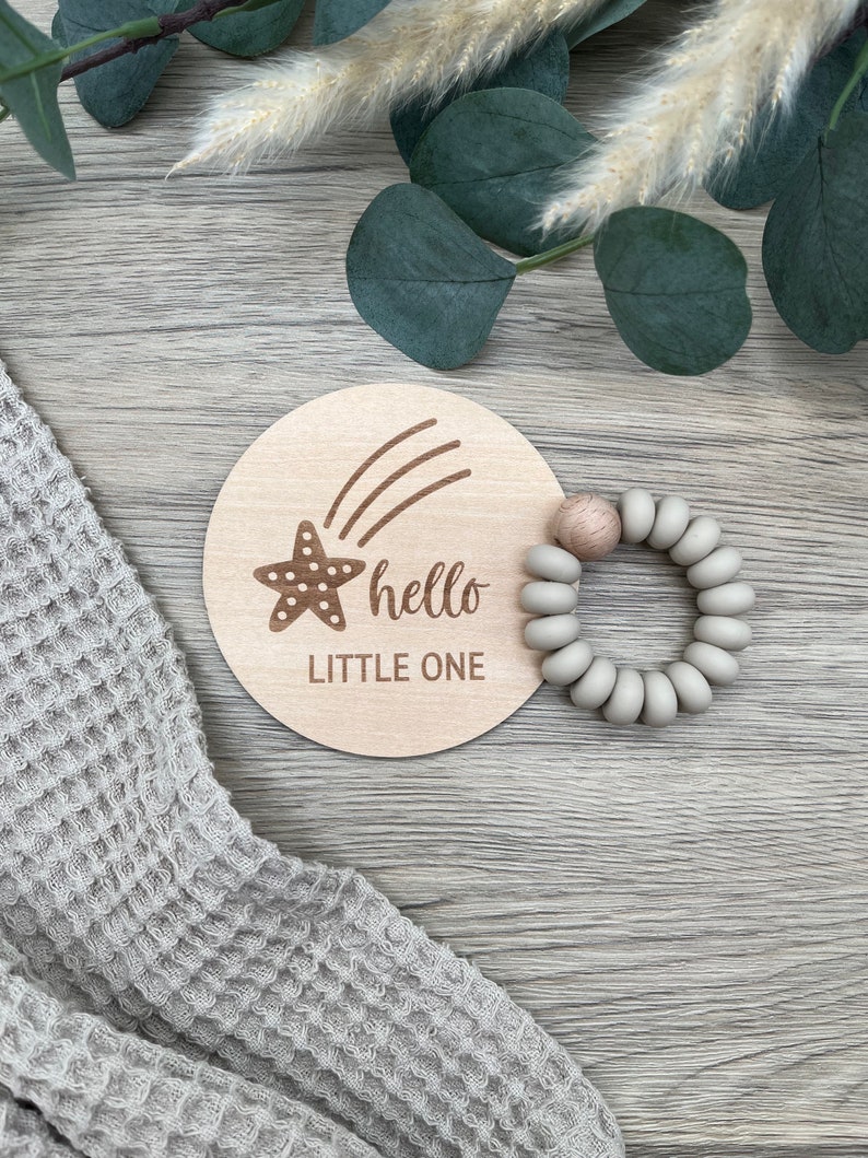 May include: A wooden round sign with a shooting star and the text "hello little one". A gray silicone teething ring is next to the sign.