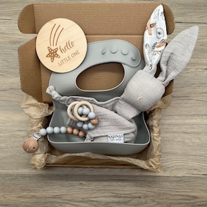 May include: A gray gift box filled with baby items, including a gray silicone bib, a wooden teether, a gray bunny rattle, and a wooden sign that says "hello little one".