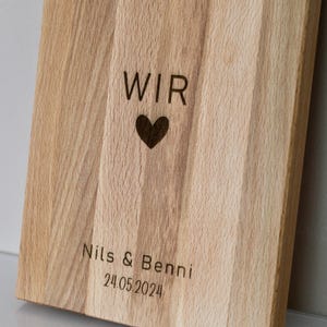 May include: A wooden cutting board with the words "WIR" and a heart engraved on it. Below the heart, it says "Nils & Benni 24.05.2024".