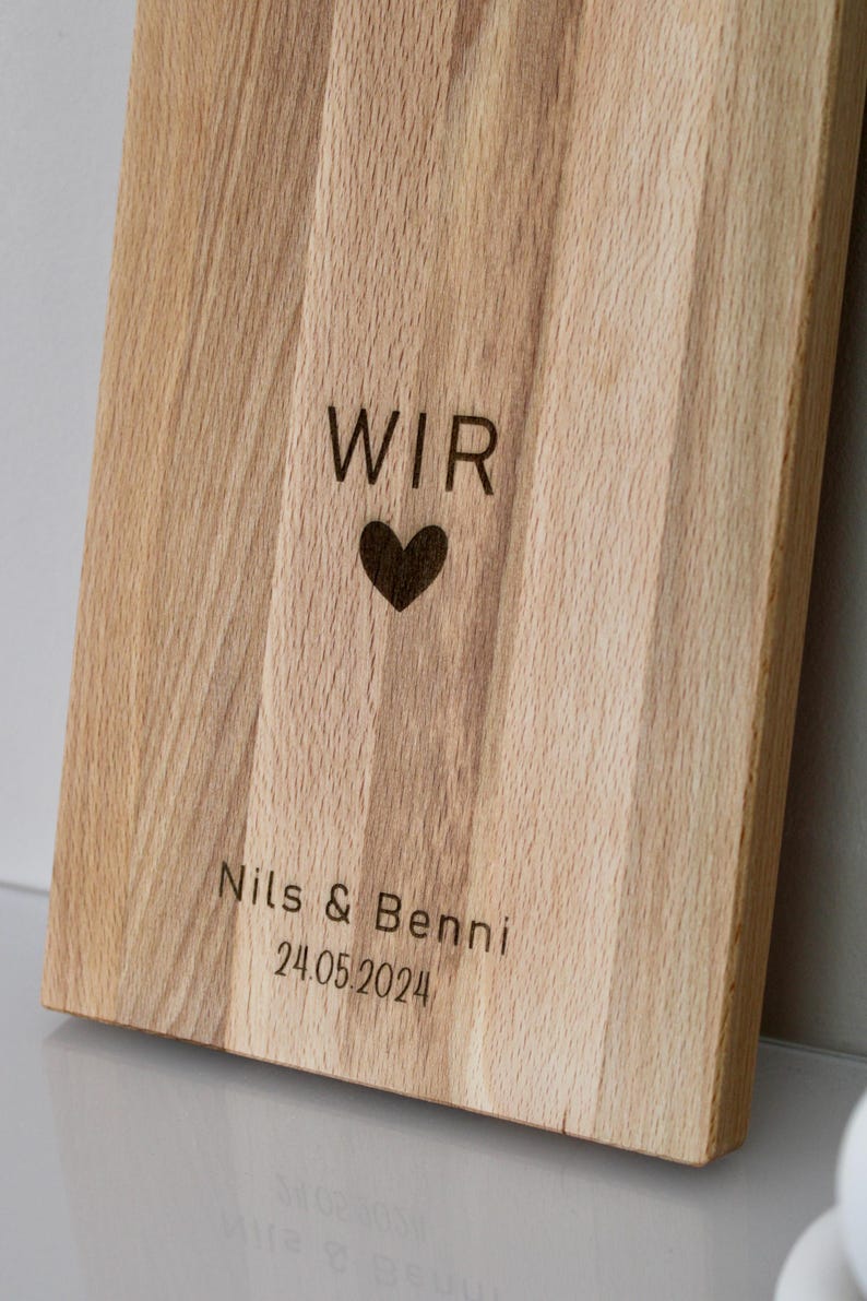 May include: A wooden cutting board with the words "WIR" and a heart engraved on it. Below the heart, it says "Nils & Benni 24.05.2024".