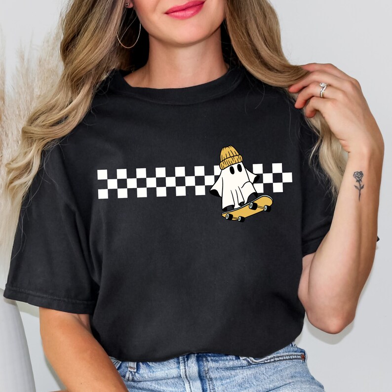 May include: Black t-shirt with a white and black checkered stripe across the chest. A white ghost wearing a yellow beanie is skateboarding on top of the stripe.