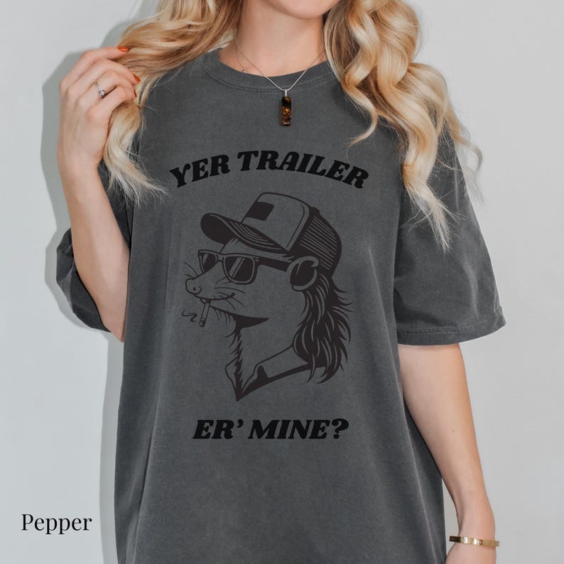 May include: Dark gray t-shirt with the text "YER TRAILER" above a graphic of a rat wearing sunglasses and a trucker hat, smoking a cigarette. Below the graphic is the text "ER' MINE?"