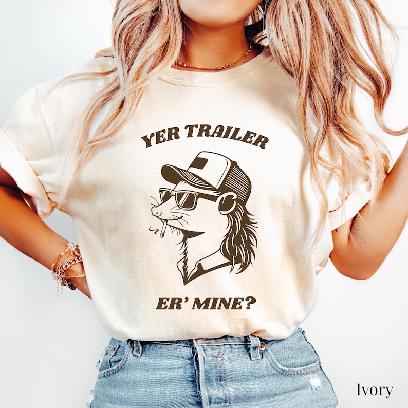 May include: Ivory-colored t-shirt with a graphic of a cartoon possum wearing sunglasses and a trucker hat, smoking a cigarette. The text "YER TRAILER ER' MINE?" is printed above and below the possum illustration. The shirt is made of a soft material.