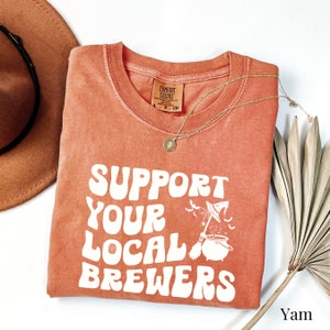 May include: A burnt orange t-shirt with the text "Support Your Local Brewers" in white. The text is in a retro font and is accompanied by a graphic of a witch's cauldron with a broom and bats.