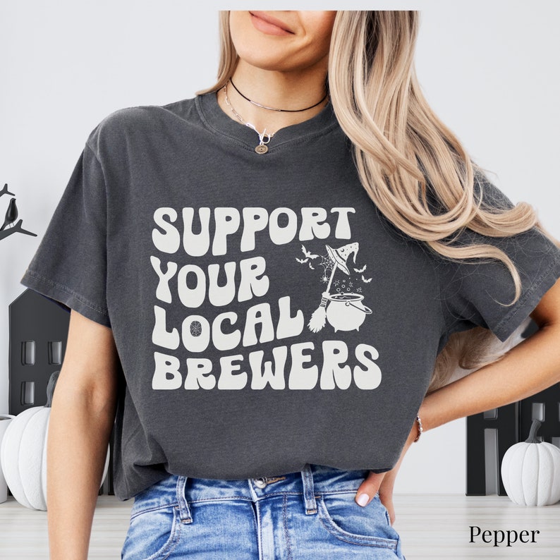 May include: A dark gray t-shirt with a white graphic that says "Support Your Local Brewers" with a witch's hat and cauldron illustration.