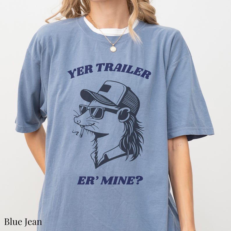 May include: Blue jean-colored t-shirt featuring a cartoon illustration of a rodent wearing sunglasses and a trucker hat, with a cigarette in its mouth. The text "YER TRAILER" is arched above the illustration, and "ER' MINE?" is below.