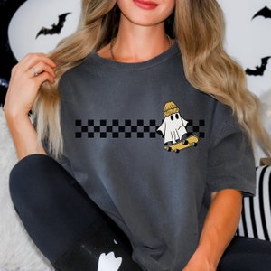 May include: A gray t-shirt with a black and white checkered stripe across the chest. A white ghost wearing a yellow beanie is skateboarding above the stripe.