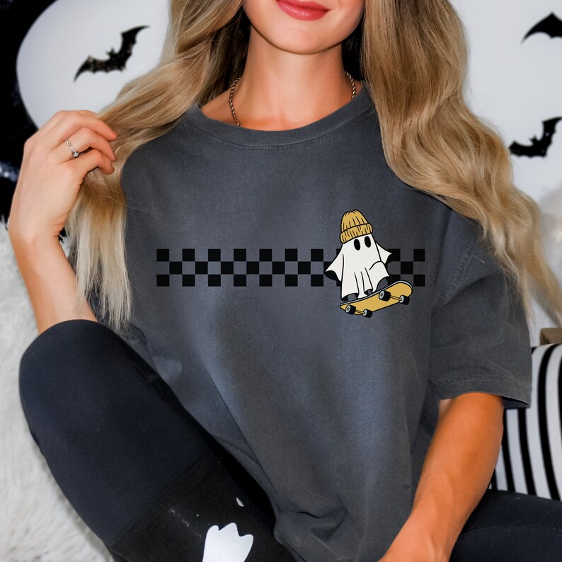 May include: A gray t-shirt with a black and white checkered stripe across the chest. A white ghost wearing a yellow beanie is skateboarding above the stripe.