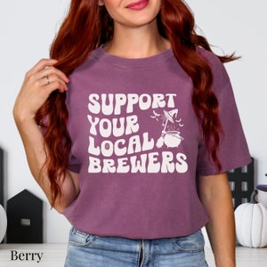 May include: A purple t-shirt with the text "Support Your Local Brewers" in white, featuring a witch's cauldron with a broom and bats.