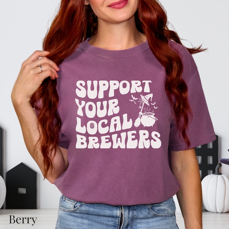 May include: A purple t-shirt with the text "Support Your Local Brewers" in white, featuring a witch's cauldron with a broom and bats.