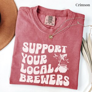 May include: A crimson red t-shirt with the text "Support Your Local Brewers" in a retro font. A witch's cauldron with a broom and bats is above the text.