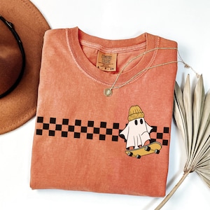 May include: A peach-colored t-shirt with a black and white checkered pattern and a ghost wearing a yellow beanie riding a skateboard. The t-shirt has a tag that says "Comfort Colors".