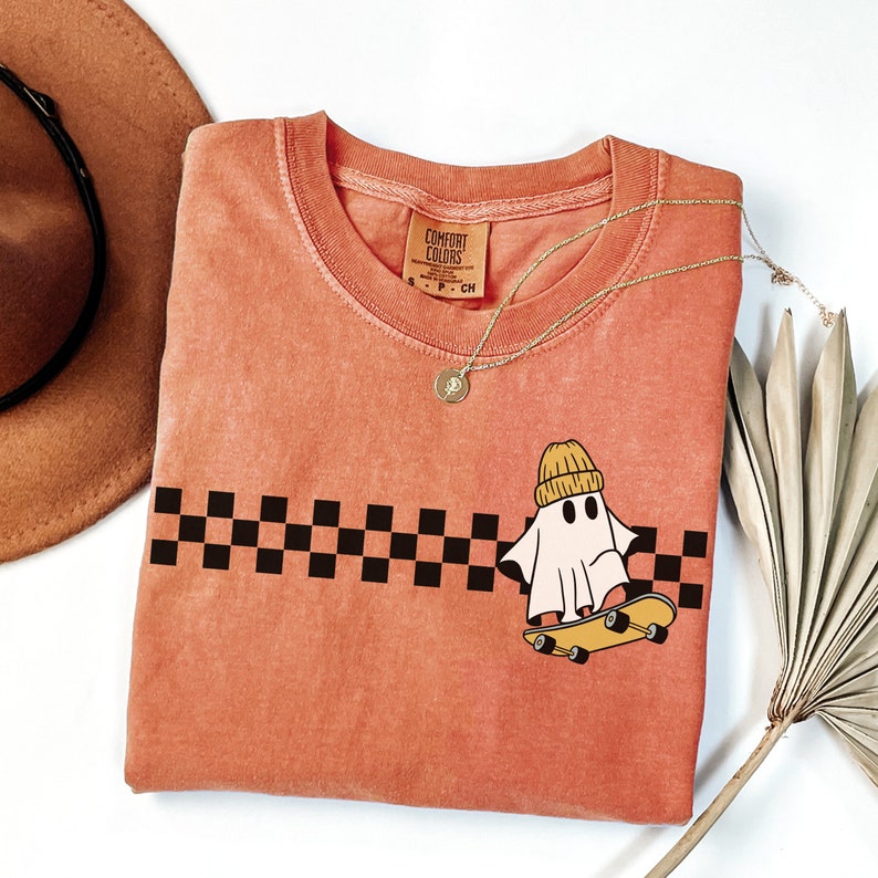 May include: A peach-colored t-shirt with a black and white checkered pattern and a ghost wearing a yellow beanie riding a skateboard. The t-shirt has a tag that says "Comfort Colors".