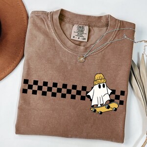 May include: A brown t-shirt with a black and white checkered pattern and a white ghost wearing a yellow beanie riding a skateboard. The text "COMFORT COLORS" is on the tag.