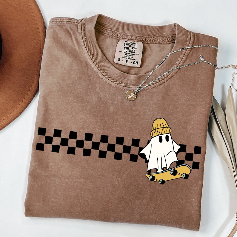 May include: A brown t-shirt with a black and white checkered pattern and a white ghost wearing a yellow beanie riding a skateboard. The text "COMFORT COLORS" is on the tag.