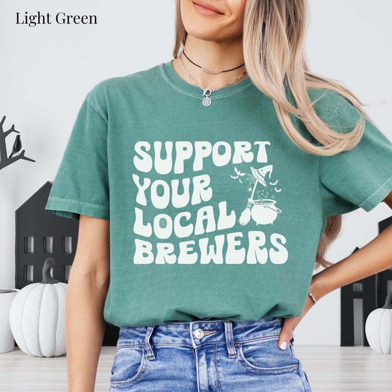 May include: A light green t-shirt with the text "Support Your Local Brewers" in white. A witch's cauldron with a broom and bats is above the text.