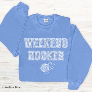 May include: A light blue sweatshirt with the text "Weekend Hooker" in white knit-like lettering. The text is surrounded by a white yarn ball and heart graphic.
