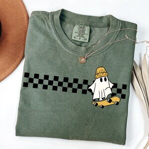 May include: A green t-shirt with a black and white checkered pattern and a cartoon ghost wearing a yellow beanie and riding a skateboard. The text "COMFORT COLORS" is on the tag.