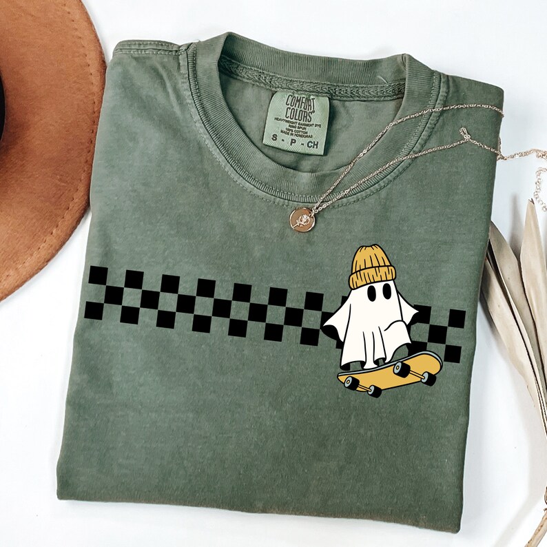 May include: A green t-shirt with a black and white checkered pattern and a cartoon ghost wearing a yellow beanie and riding a skateboard. The text "COMFORT COLORS" is on the tag.