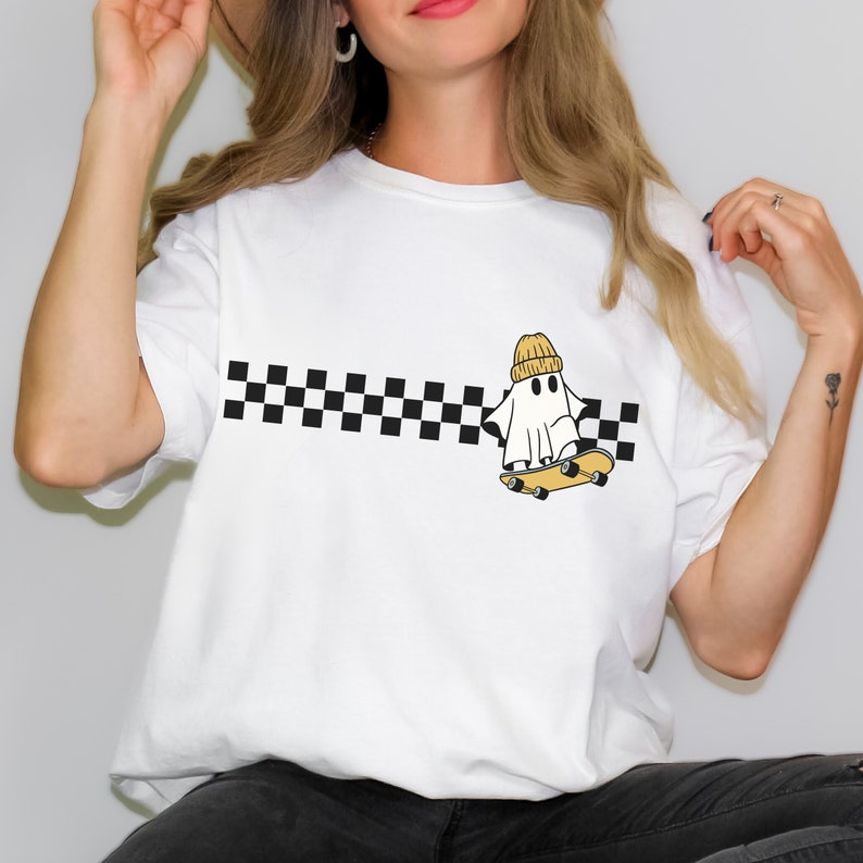 May include: White t-shirt with a black and white checkered stripe across the chest. A cartoon ghost wearing a yellow beanie is skateboarding.