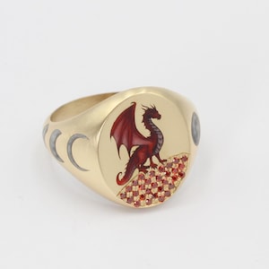 May include: A gold signet ring with a red dragon design and a crescent moon pattern on the band. The dragon is set against a background of red gemstones.