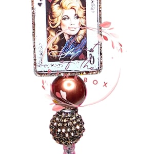 Könnte beinhalten: Dekorativer Flaschenverschluss mit einem Queen of Hearts-Spielkarten-Design mit einem Porträt von Dolly Parton. Der Verschluss hat eine braune, juwelenbesetzte Basis und eine rosa-braune dekorative Oberseite.