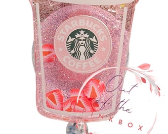 May include: A pink glittery Starbucks coffee cup badge reel featuring a white lid, a green straw, and embedded strawberry slices. The center displays the circular Starbucks Coffee logo. A metal clip is attached to the bottom of the cup.