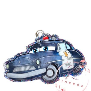 May include: A blue and silver police car keychain with a red light on top. The car has large blue eyes and the word "SHERIFF" on the side. The keychain is outlined with iridescent glitter.