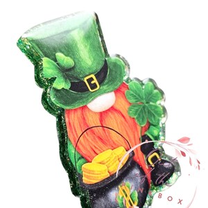 May include: A green leprechaun gnome with a pot of gold coins. The gnome is wearing a green hat with a gold buckle and a green shirt. The gnome is holding a pot of gold coins in one hand and a shamrock in the other.