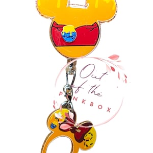 May include: A yellow and red badge reel featuring a Mickey Mouse head design with a honey pot graphic. The badge reel has a dangling charm of a Mickey Mouse ear headband. The text "Out of the Pink Box" is in a pink circle.