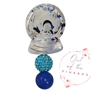 May include: A clear snow globe charm with a penguin inside. The globe is attached to a blue and turquoise beaded chain. The charm is labeled "Out of the Pink Box".