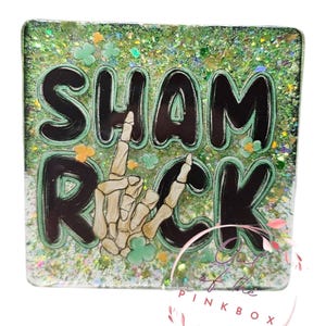 May include: A square, glittery green resin item with the words "SHAM ROCK" in black, outlined in green. A skeleton hand makes a rock-and-roll gesture. The background is filled with glitter and shamrock shapes.