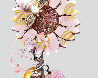 Puede incluir: Un carrete de credencial transparente con un dije de girasol rosa y dorado, adornado con una abeja. El carrete de credencial está decorado con cuentas rosas y doradas y un dije que dice "Out of the Pink Box".