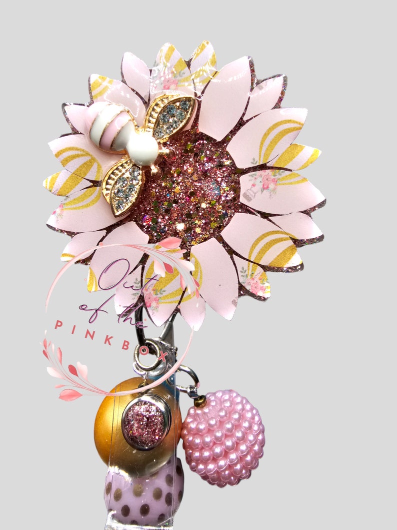 Puede incluir: Un portacredenciales con un girasol rosa y dorado con centro brillante y un detalle de abeja. Bajo la flor, un clip met&aacute;lico sostiene una cuenta dorada, una cuenta rosa brillante y un dije de cuentas rosa. El texto Out of the PINK BOX es visible.
