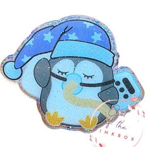 May include: A light blue penguin-shaped accessory with a glittery finish. The penguin wears a blue nightcap with star patterns and holds a video game controller. The design includes closed eyes, a pacifier, and yellow feet. The text "Out of the Ink Box" is visible.