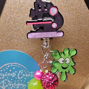 May include: A black and pink microscope charm with a green slime character charm, pink and green beads, and a pink rhinestone charm. The charms are attached to a silver chain.