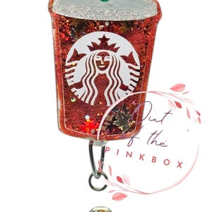 May include: A red glittery badge reel shaped like a coffee cup, featuring the Starbucks logo in white. The cup has a silver glitter rim and a green straw. A matching red glitter button is attached.