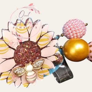 Puede incluir: Un portatarjetas con forma de girasol con p&eacute;talos de color rosa claro, rayas doradas y un centro de color rosa oscuro brillante. Un peque&ntilde;o dije de abeja con pedrer&iacute;a transparente se asienta sobre los p&eacute;talos. Incluye tres cuentas: una cuenta rosa texturizada, una cuenta dorada lisa y una cuenta rosa con lunares dorados.