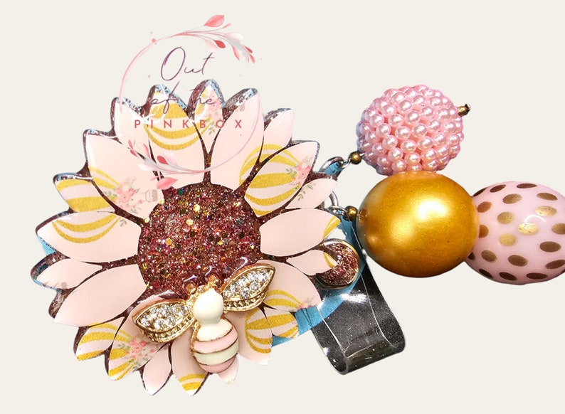 Puede incluir: Un portatarjetas con forma de girasol con p&eacute;talos de color rosa claro, rayas doradas y un centro de color rosa oscuro brillante. Un peque&ntilde;o dije de abeja con pedrer&iacute;a transparente se asienta sobre los p&eacute;talos. Incluye tres cuentas: una cuenta rosa texturizada, una cuenta dorada lisa y una cuenta rosa con lunares dorados.