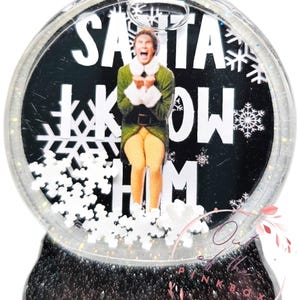 May include: A snow globe with a black background and white snowflakes. The globe features a character from the movie Elf, Buddy the Elf, with the text "Santa Know Him".