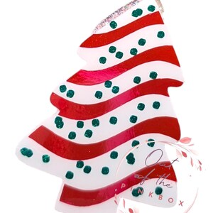 May include: A white Christmas tree bauble with red and white stripes and green polka dots. The bauble is decorated with glitter and has a shiny finish. The bauble is labelled "Out of the Pinkbox".