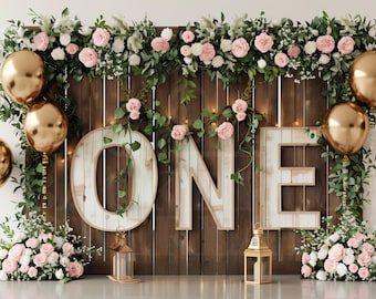 May include: A wooden backdrop decorated with greenery, pink flowers, and gold balloons. The backdrop features the word "ONE" in large, rustic letters. Two lanterns are placed on the ground in front of the backdrop.
