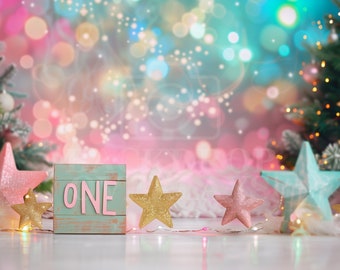 May include: A light blue wooden block with the word "ONE" in pink letters. The block is sitting on a white surface in front of two Christmas trees decorated with lights and stars. The background is a blurred image of pink, blue, and green lights.