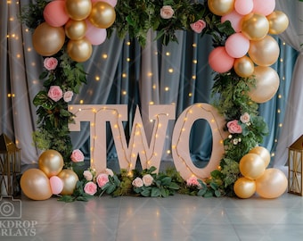 May include: Two large wooden letters spelling "TWO" decorated with lights and greenery. The letters are surrounded by pink and gold balloons and flowers.