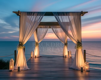 May include: A wooden wedding archway draped with white sheer fabric, decorated with greenery and lanterns, stands on a wooden dock overlooking a calm ocean at sunset.