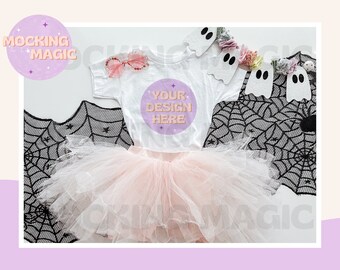 May include: A white t-shirt with the text "YOUR DESIGN HERE" in a pink circle. The shirt is on a white surface with a black spiderweb, a pink tulle skirt, and a string of white ghosts with rainbow yarn.