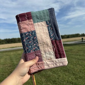 Puede incluir: Una funda de libro de patchwork en tonos rosa, azul, verde y burdeos. La funda tiene un estampado floral y una costura decorativa. Se adjunta un marcapáginas de cinta amarilla y marrón.